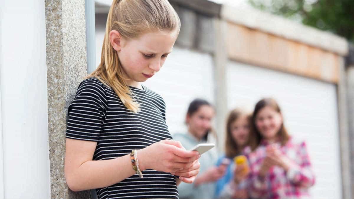 pre-teen girl looks at cell phone as schoolmates laugh