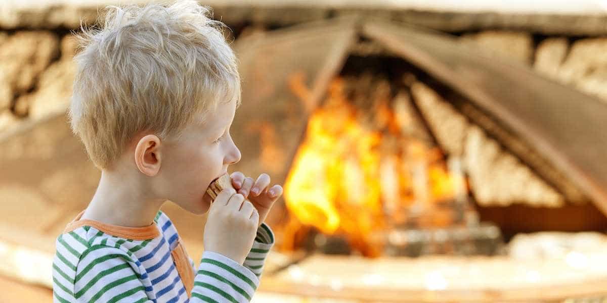 Boy eating s'mores in front of campfire and tent at campsite