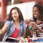 teenage high school girls with chemistry equipment and laptop at home